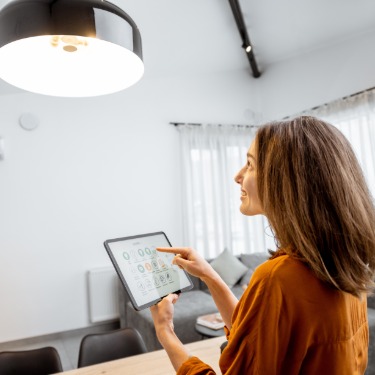 Woman controls a ceiling light with a tablet displaying smart home settings in a bright, modern living room.