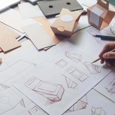 A designer's hand using a pencil to sketch an idea for a new product's packaging on multiple sheets of paper.