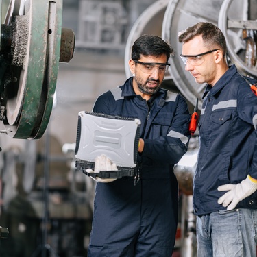 Two technicians look at a laptop while standing between machines in a plant. They wear dark blue jackets and jumpsuits.