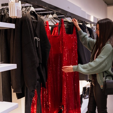Woman in a clothing store examines a bright red sequined dress hanging among black garments on a rack.