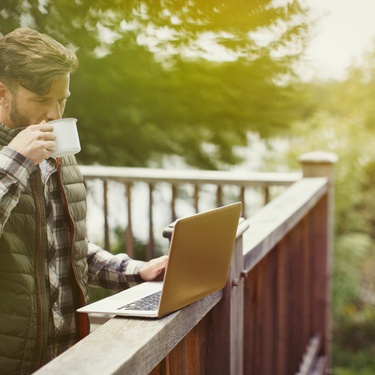A man drinking coffee and looking at his laptop while standing on the deck of his cabin. Trees stand in the background.