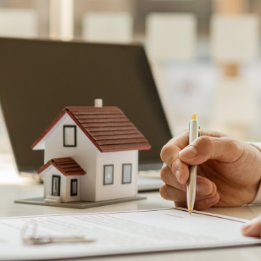 A close-up of a hand holding a pen over a document on a desk, next to a small, white model house and a laptop.