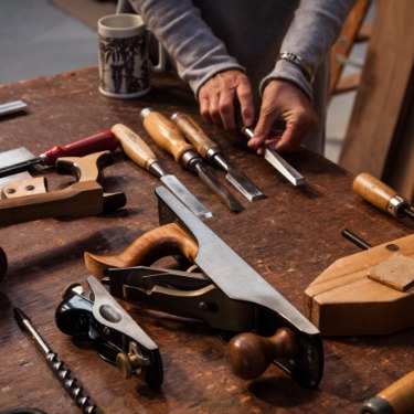 A man is placing a chisel on a wooden table. The table is filled with other woodworking tools.