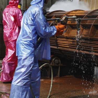 A person wearing a blue protective suit and a person wearing a pink protective suit are cleaning an industrial machine.