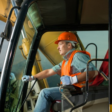 A heavy equipment operator wearing a hard hat, safety vest, and gloves while sitting at the controls of a machine.