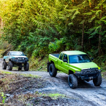 Two lifted pickup trucks with oversize off-road tires drive along a rugged dirt road surrounded by dense forest.