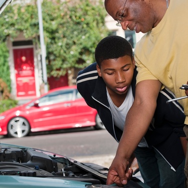 A father smiles as he shows his son the parts of a car engine. A red car sits parked in the street behind them.