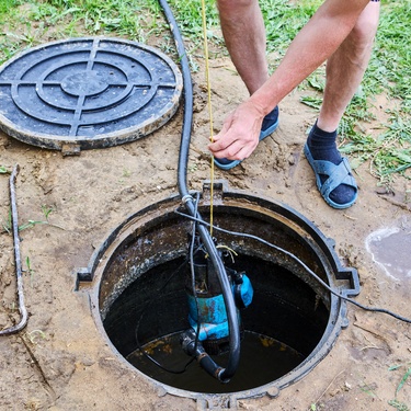 A person is removing a submersible pump from a private well with a flexible drop pipe. A manhole cover is lying in the dirt.