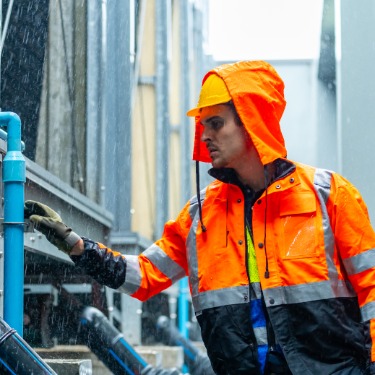 Industrial worker wearing a bright orange high-visibility rain jacket and hard hat inspects equipment and blue pipes outdoors during heavy rainfall.