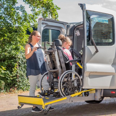 A woman stands behind a full-size van with the back doors open and a wheelchair lift deployed. A girl in a wheelchair is on the lift.