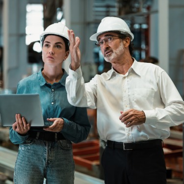 A man and a woman wearing hard hats stand on the production floor of an industrial facility. The man raises his hand.