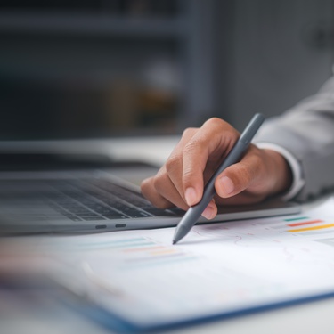 A person reviewing paperwork on a clipboard with financial graphs on it. Their laptop is sitting next to the clipboard.