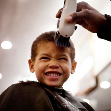 A boy wearing a salon cape getting a haircut with electric clippers while seated and smiling with his eyes closed.