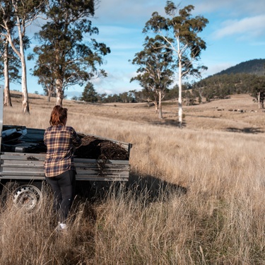 A person standing beside a white utility truck in a dry grassy field with trees and low hills in the background.