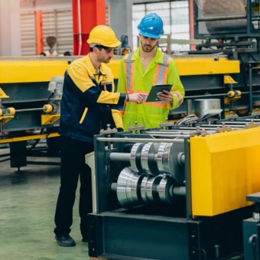 Two employees wearing hard hats in a brightly lit manufacturing facility. Both employees look at a tablet.