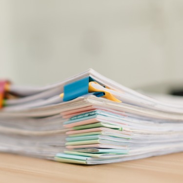 A large pile of white documents next to a small laptop on a wooden desk in a brightly lit office space.
