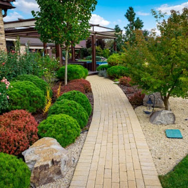 A winding white brick path curves through colorful landscaped shrubs and mature trees beside a well-kept home.