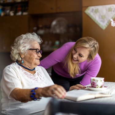 A young woman smiles while spending time with her elderly female family memeber at a table in a warm home setting.