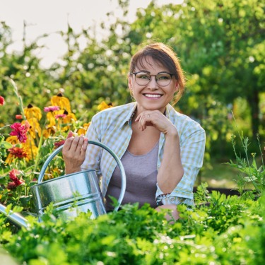 A smiling woman gardener in a plaid shirt holds a watering can among flowers and green plants in a lush garden.