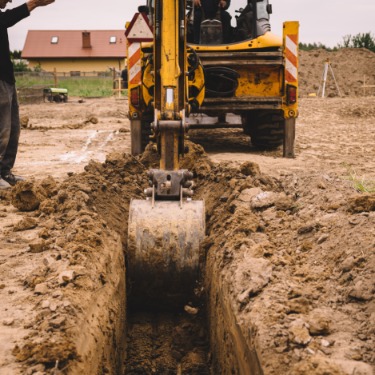A yellow excavator digs a trench in muddy ground at a construction site with a worker standing nearby.