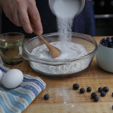 A person pouring sugar into a bowl of flour. A bowl of blueberries sits next to the flour and sugar.