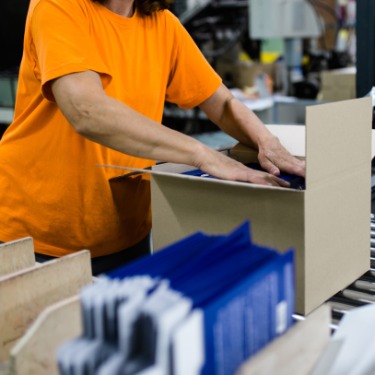 Someone stands in front of an open cardboard box on a roller conveyor. A stack of products is blurred in front of the box.