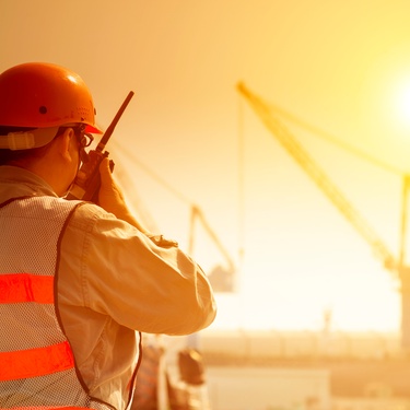 A worker in safety gear speaking into a radio at a sunlit construction site with cranes in the background.