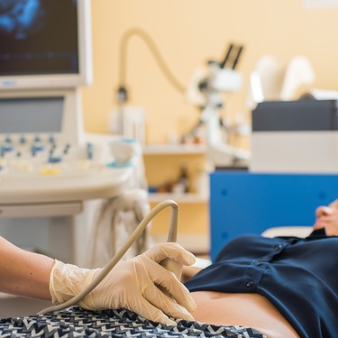 A person wearing gloves moves an ultrasound probe across a pregnant woman's abdomen beside medical equipment.