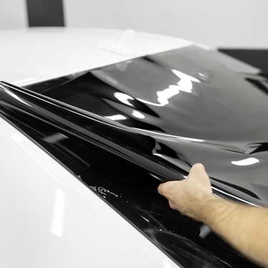 A person placing a dark black window tint on the back window of a white car inside a brightly lit room.