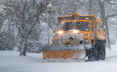 A large, yellow snowplow drives down a residential street, pushing the snow off the road. Snow continues to fall.